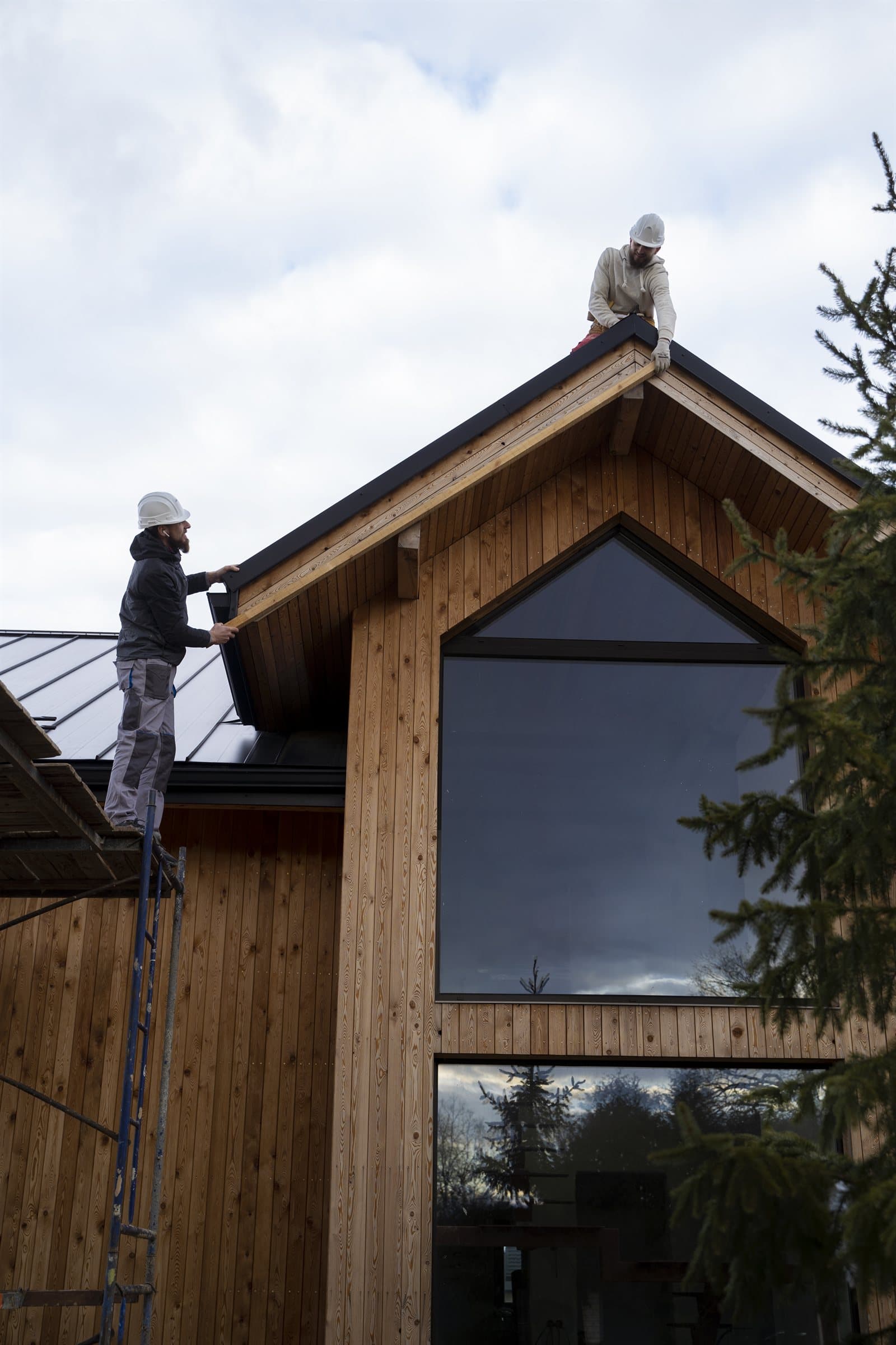 Two Optik roofers in hard hats fitting trim along the roofline of a timber-clad home